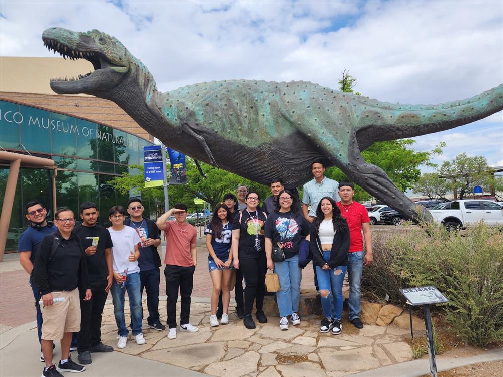 A group of people poses in front of a large dinosaur sculpture outside a museum, with a cloudy sky in the background.