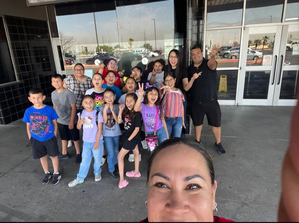 A group of children and adults pose for a photo outside a building, with a woman in the foreground taking a selfie.