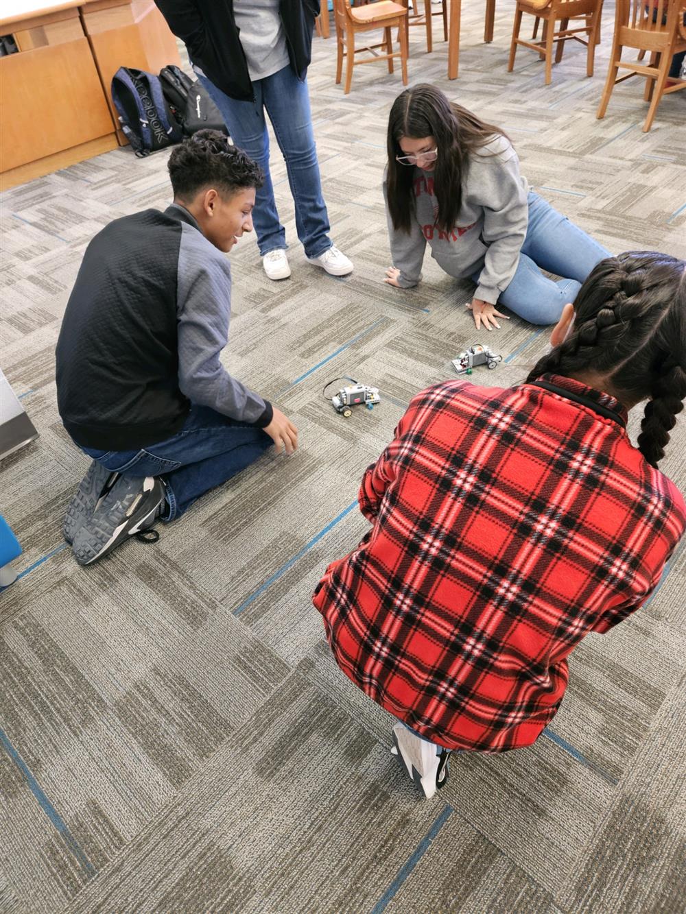 A group of four students is gathered on the floor of a library, focused on small robotic devices in front of them, with two students kneeling and two sitting nearby.