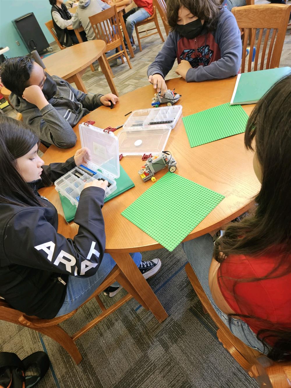 A group of children are gathered around a table in a library, engaged in a hands-on activity involving building or programming with robotics kits, with green sheets of paper spread out on the table.