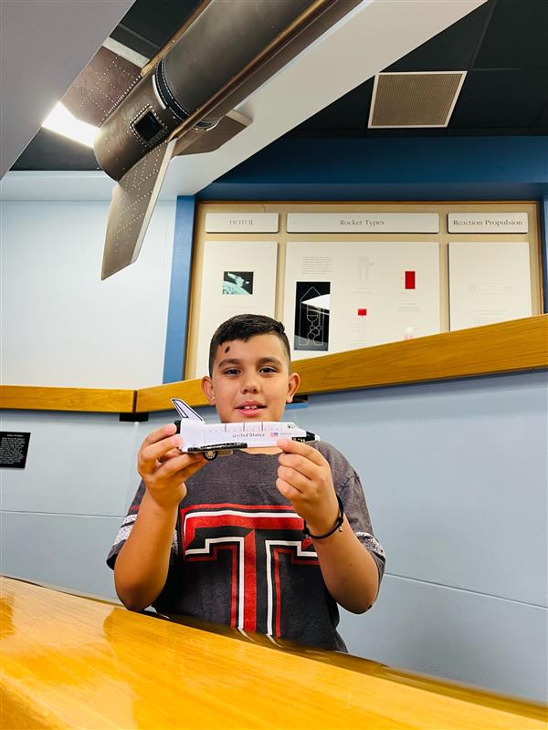 A young boy holds a model airplane while sitting at a wooden table in a room with a model rocket displayed above him.
