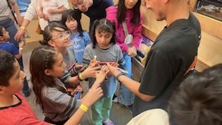 A group of children eagerly gather around an adult, reaching out to touch or hold an object being presented, in a lively educational setting.