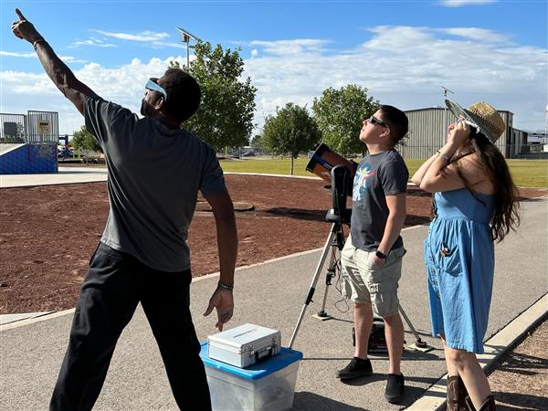 Three people outdoors, one pointing towards the sky while the others look up, with a blue sky and clouds in the background.