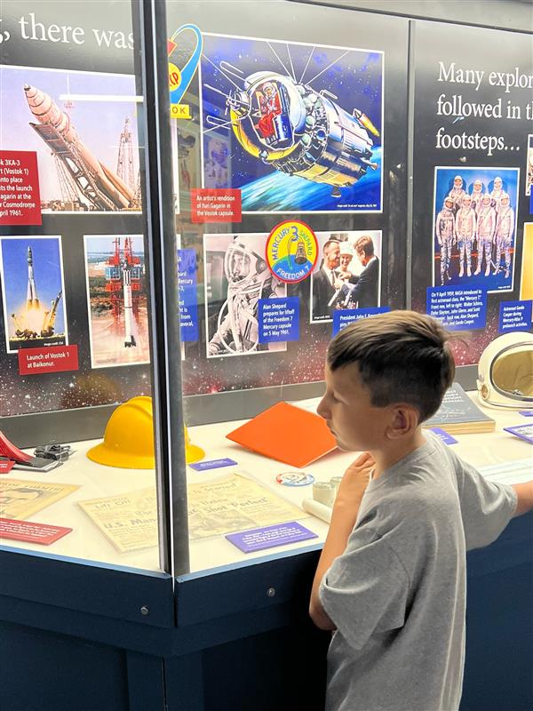 A young boy observes a display case featuring various space exploration artifacts and images, including rockets and astronaut gear, in a museum setting.