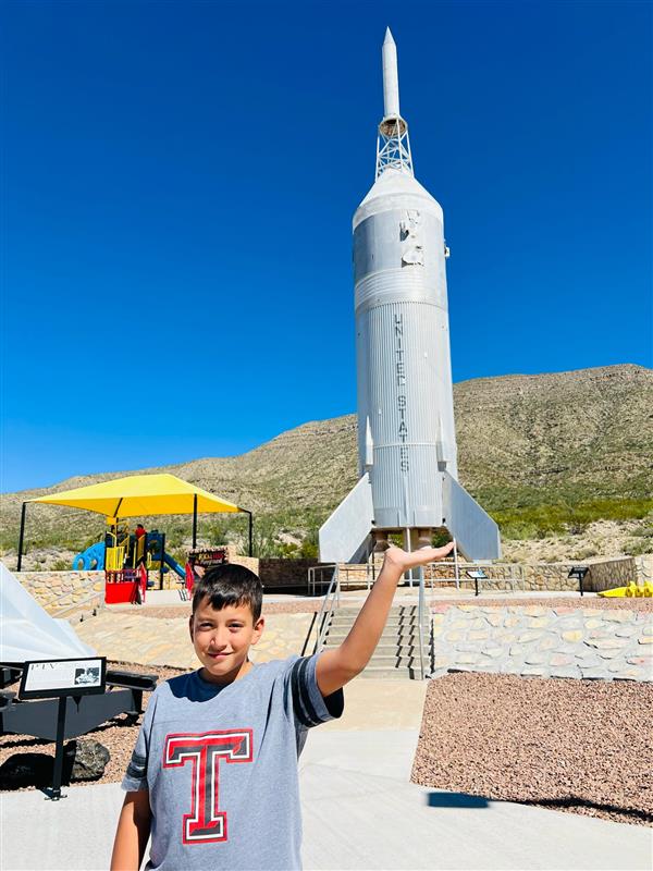 A young boy stands outdoors, smiling while holding a model rocket above his head, with a clear blue sky and mountains in the background.