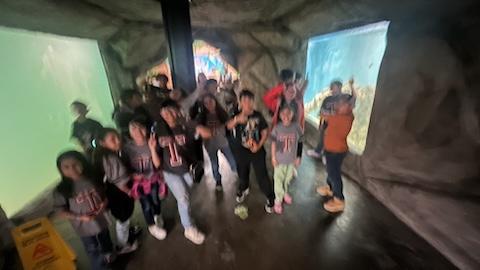 A group of children and adults are gathered in an aquarium tunnel, posing for a photo with large tanks of water on either side, showcasing aquatic life.