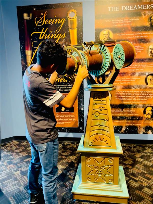A person is looking through a large, ornate telescope-like device in a museum setting, with educational posters in the background.