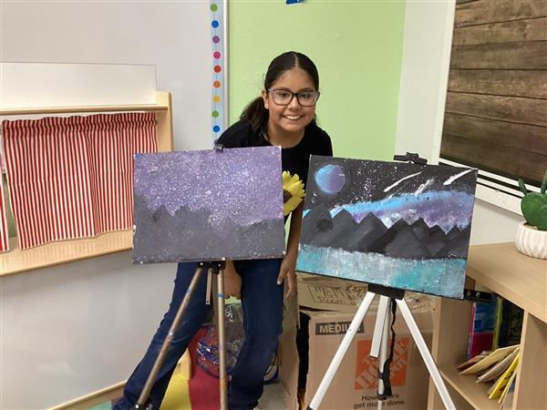 A young girl wearing glasses smiles while standing between two paintings on easels, showcasing her artwork featuring a starry night and mountain landscape.