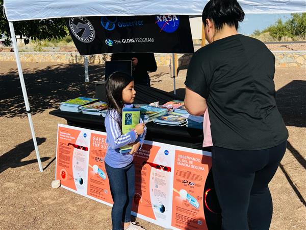 A young girl in a blue jacket is receiving materials from an adult woman at a booth under a white tent, with informational posters displayed on the table.