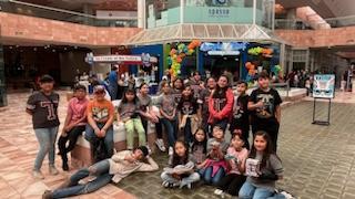 A large group of children and teenagers poses together in a mall atrium, with colorful balloons and decorations in the background.