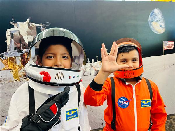Two children dressed as astronauts pose in front of a space-themed backdrop, one wearing a helmet and the other making a hand gesture, with a lunar landscape and Earth visible behind them.