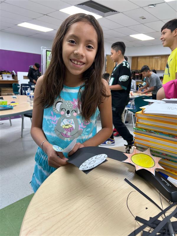 A young girl with long hair smiles while holding a craft project in a classroom filled with students and school supplies.