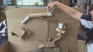A person is demonstrating a homemade contraption made from cardboard and tubes, showcasing a simple machine or Rube Goldberg device.