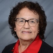 A woman with curly hair, wearing glasses and a red shirt under a black jacket, smiles at the camera against a textured gray background.