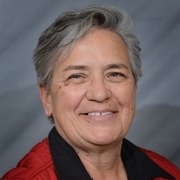 A woman with short hair wearing a black blazer over a red shirt, standing in front of a textured gray background.