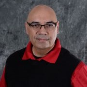 A middle-aged man with glasses, wearing a black vest over a red shirt, poses against a textured gray background.