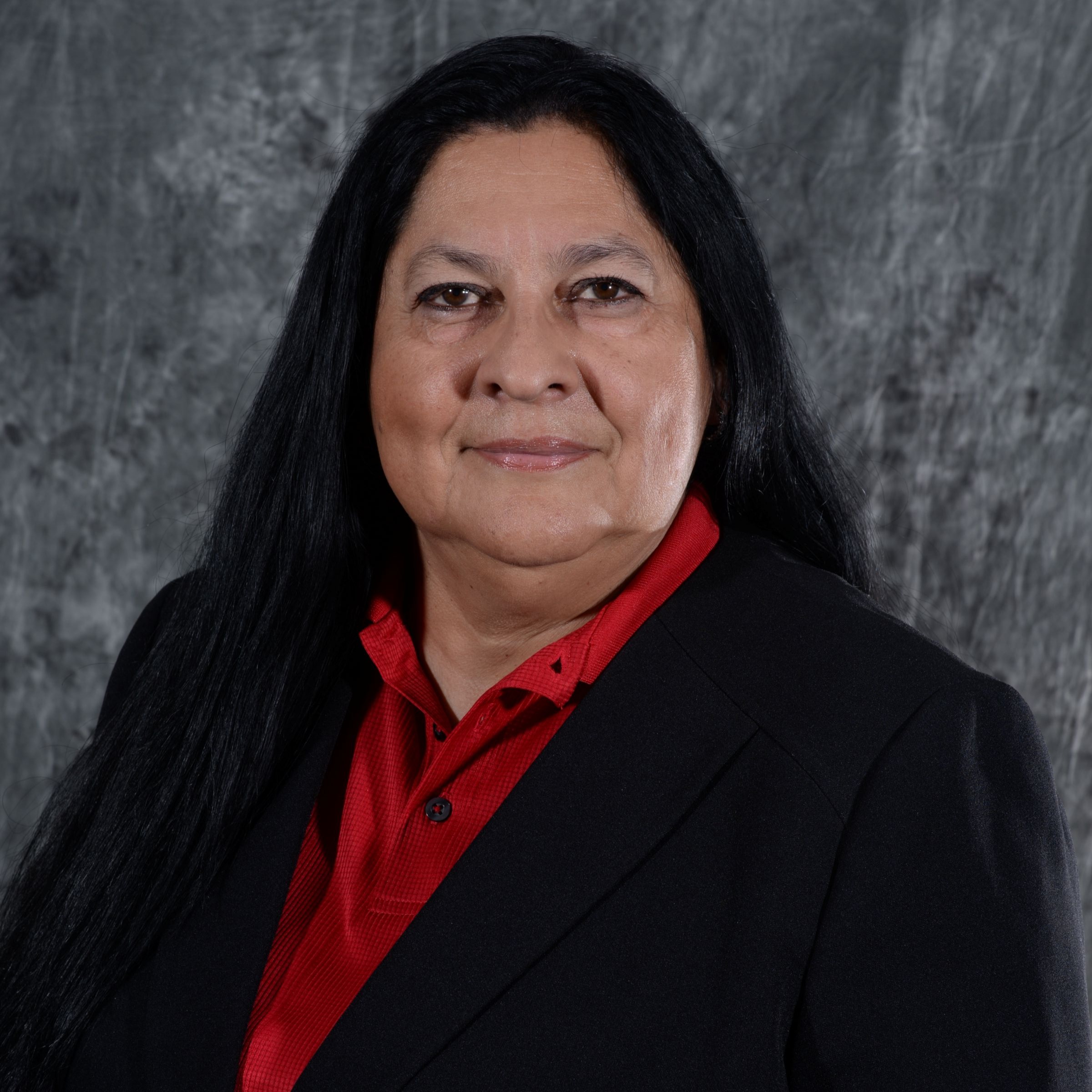 A woman with long black hair wearing a black suit and a red blouse stands in front of a textured gray background, smiling softly at the camera.