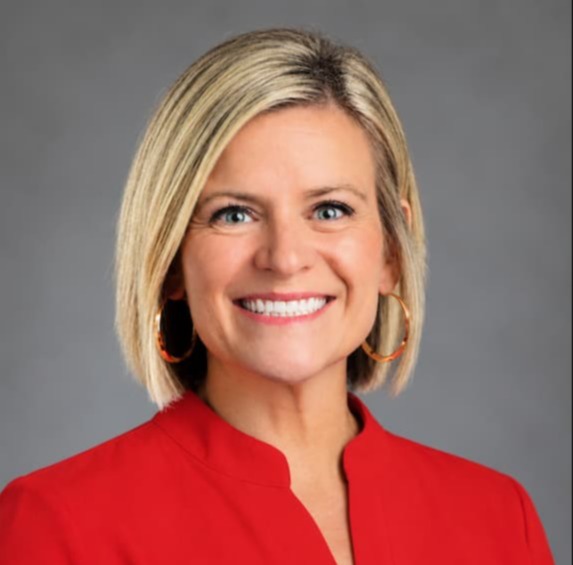 A woman with short blonde hair, wearing a red top and smiling. She has hoop earrings and a bright smile.