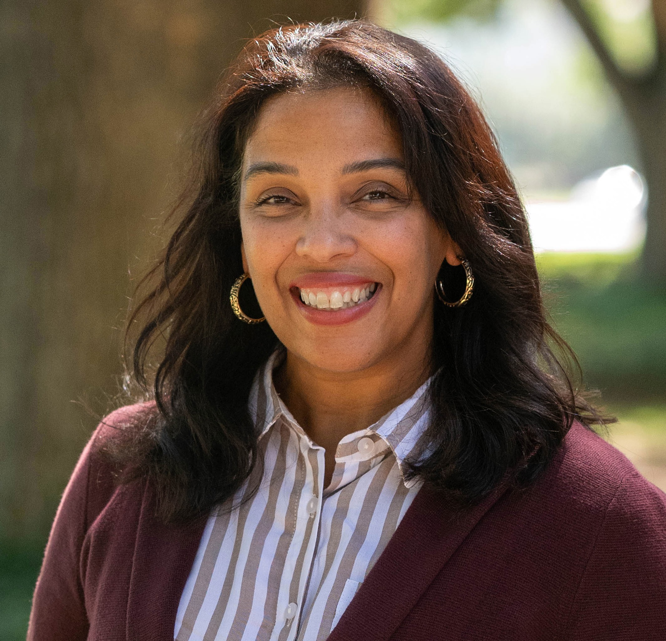 A woman wearing a striped shirt and a maroon cardigan smiles in front of a tree.