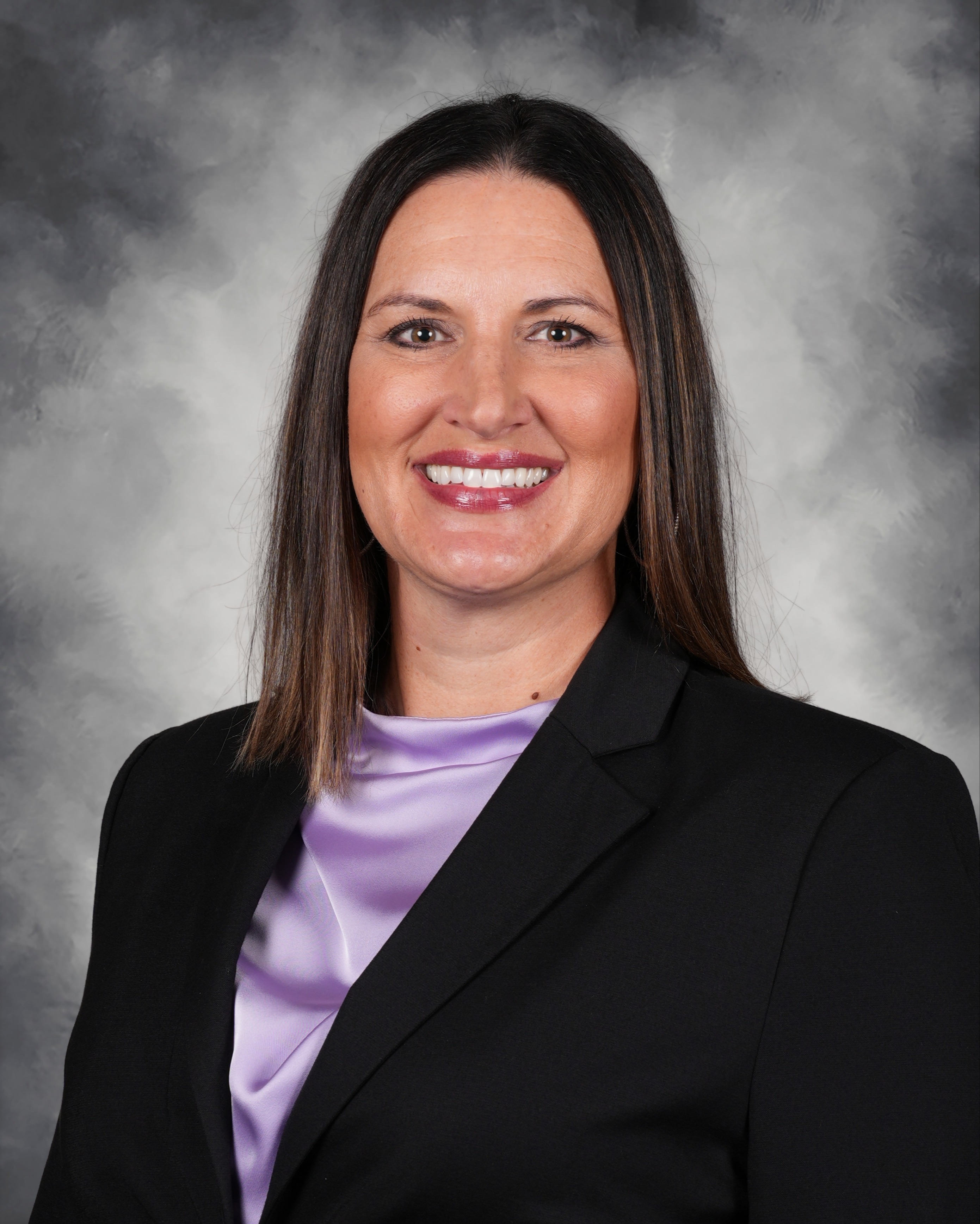 A woman in a black blazer and purple blouse smiles for a photo against a blurred gray background.