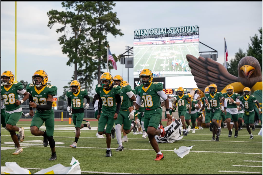 Football players in green uniforms and helmets run on a field. A large mascot and scoreboard are in the background.