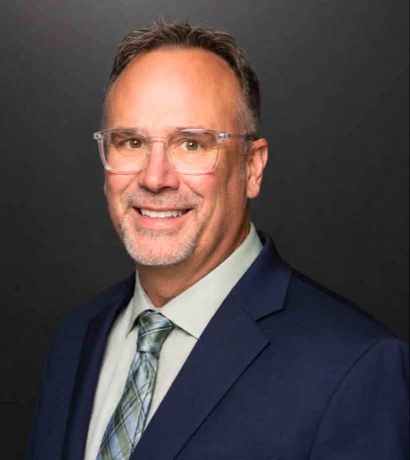 A man with short hair, a trimmed beard, and glasses smiles. He wears a suit and tie.
