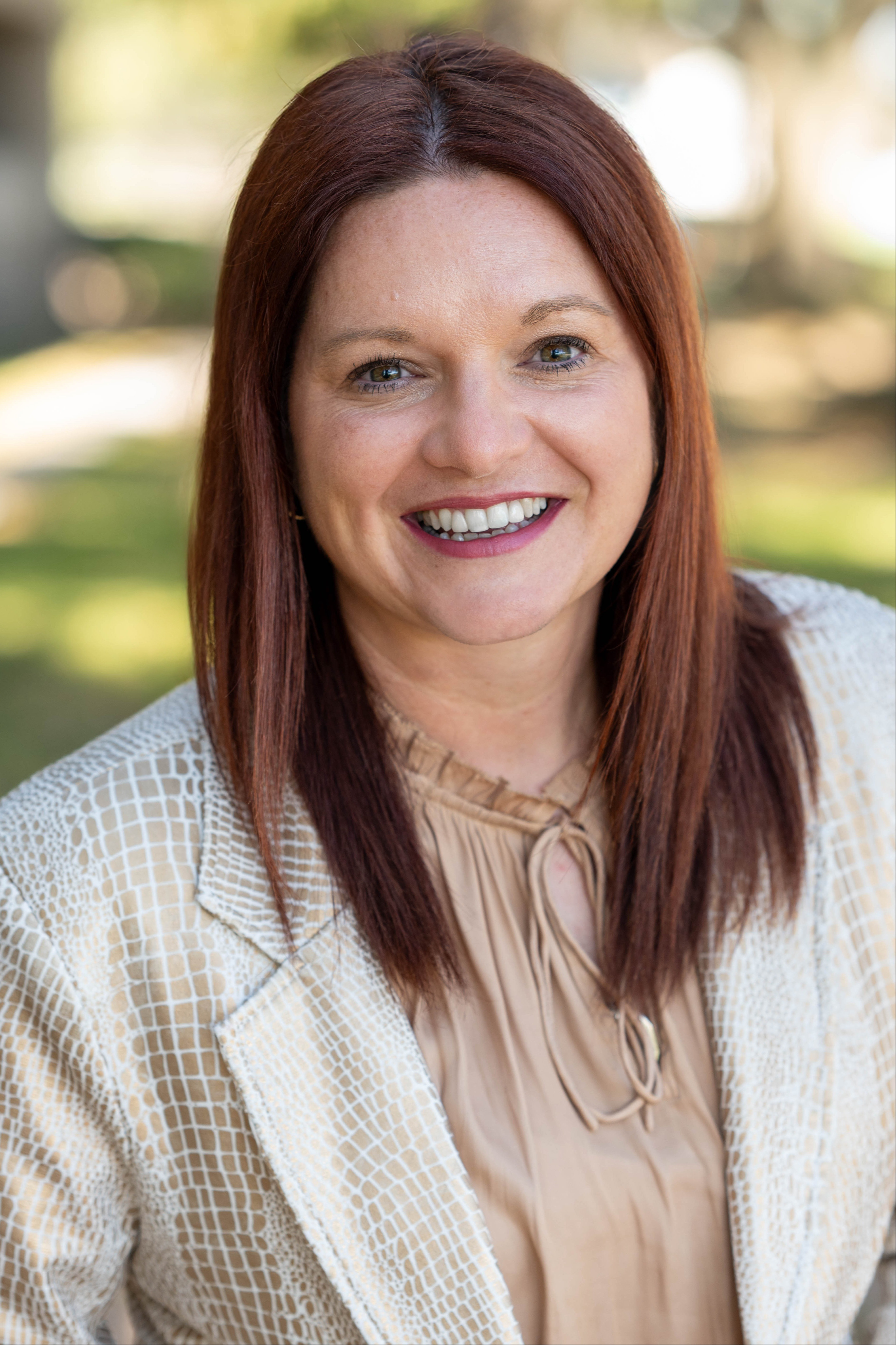 A woman with red hair wearing a light-colored blazer and blouse stands outside with a smile.