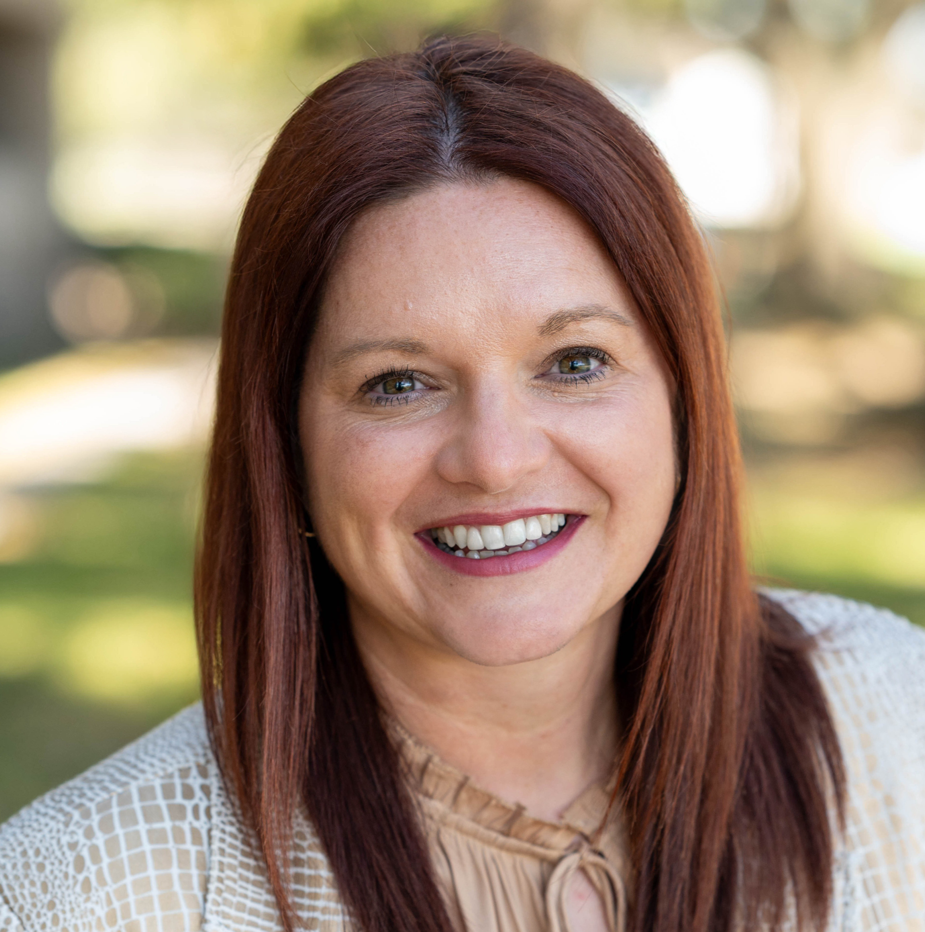 A woman with red hair wearing a light-colored blazer and blouse stands outside with a smile.