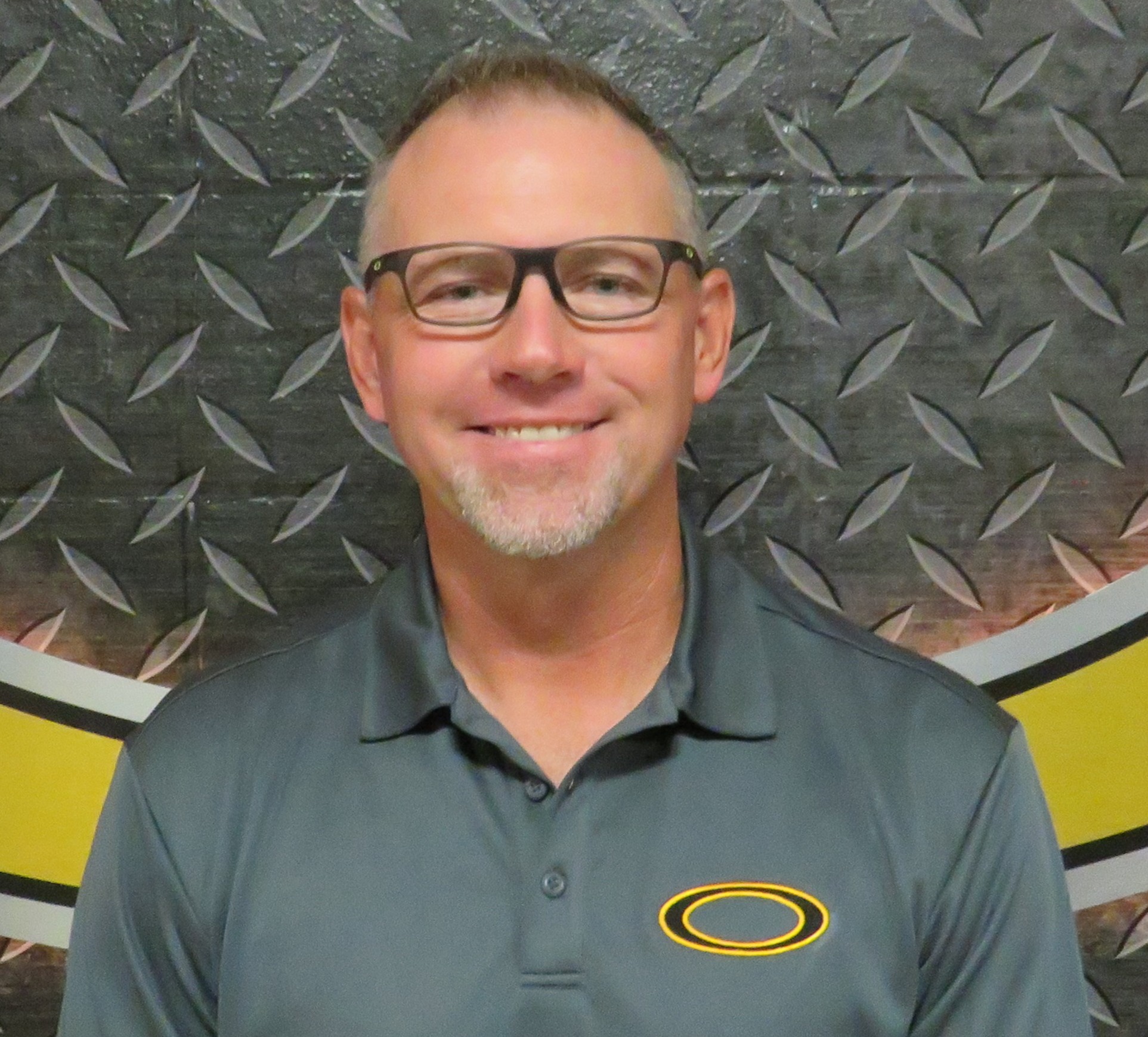 Man in a gray polo with a yellow logo, wearing glasses, smiling, standing in front of a yellow and gray wall.