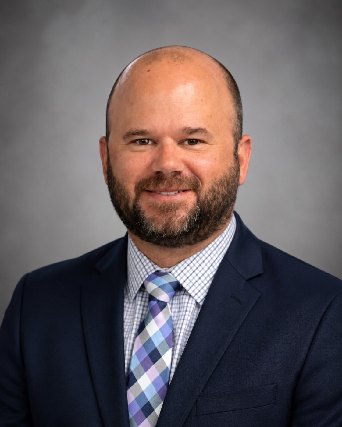 Man with beard, wearing navy suit, blue and white checkered shirt, and colorful tie, against gray background.