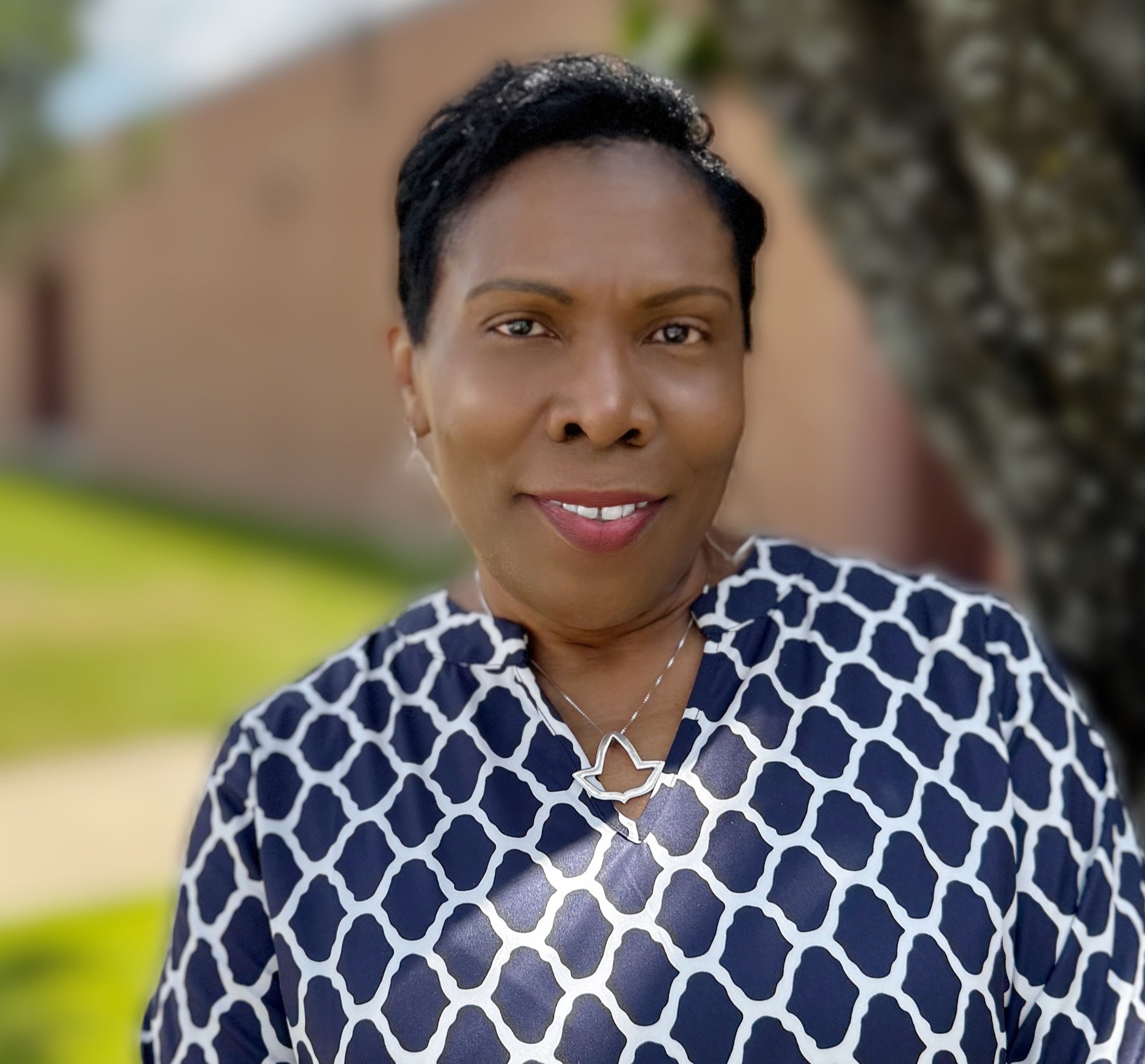A woman wearing a blue and white patterned shirt stands with a smile in front of a tree.