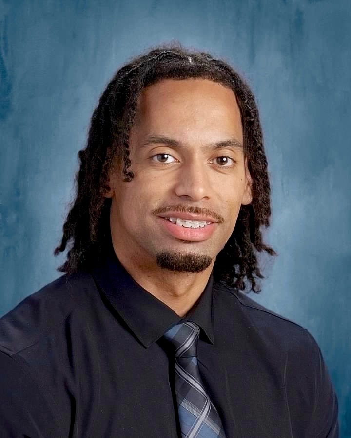 Man with short dreadlocks wearing a black shirt and a blue tie. He is smiling.