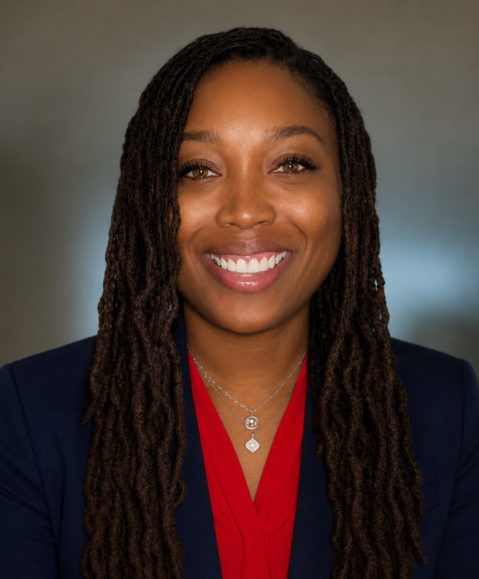 Woman with curly hair smiling at camera, wearing a red top and blue blazer with a silver necklace.