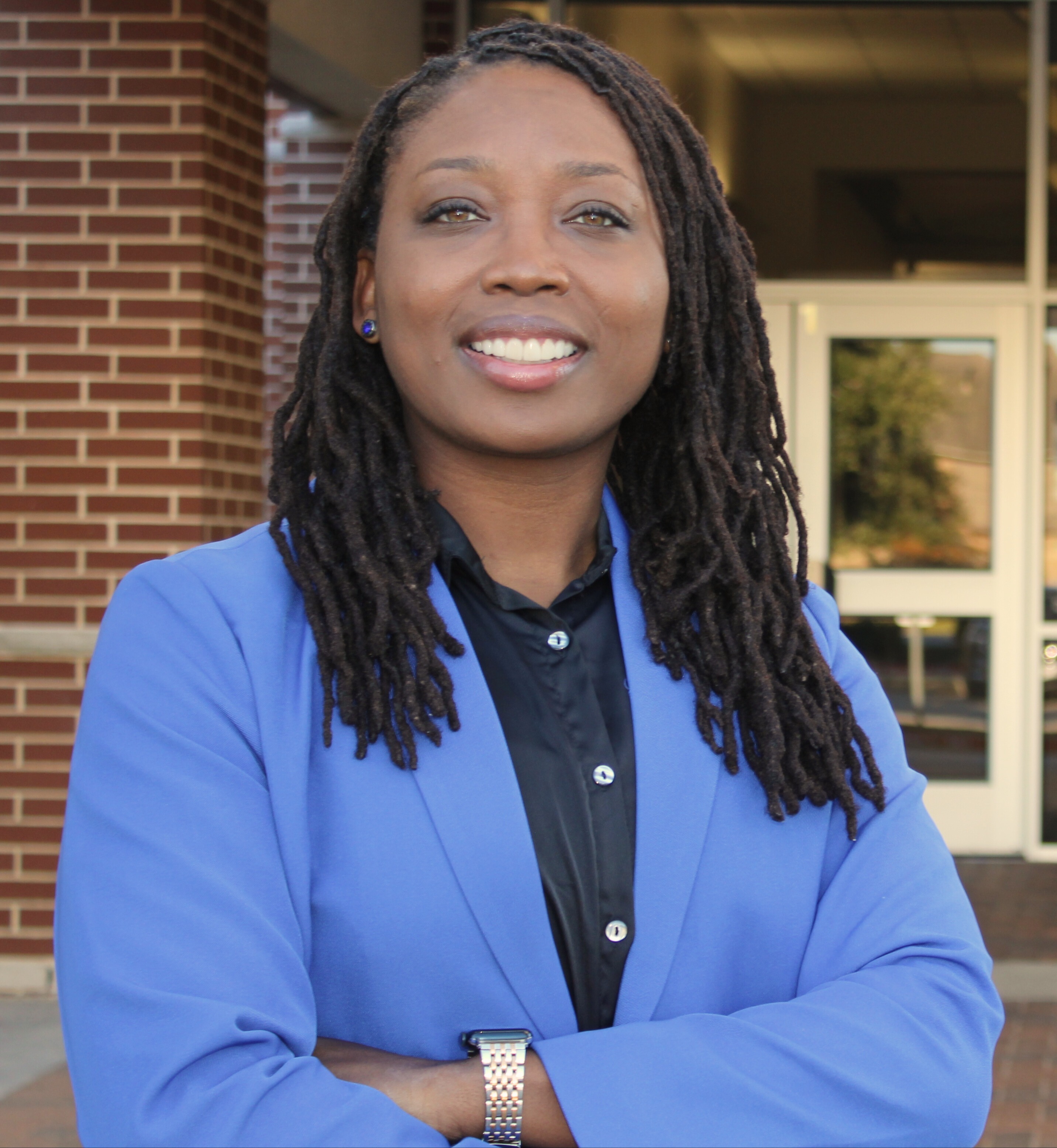 Woman with braided hair wearing a blue jacket and black top standing with arms crossed outside a brick building.