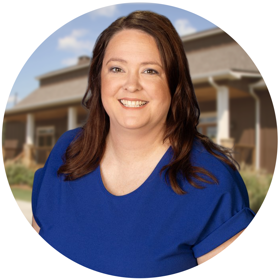 A woman with long brown hair, wearing a blue shirt, smiles in front of a building with a porch in the background.