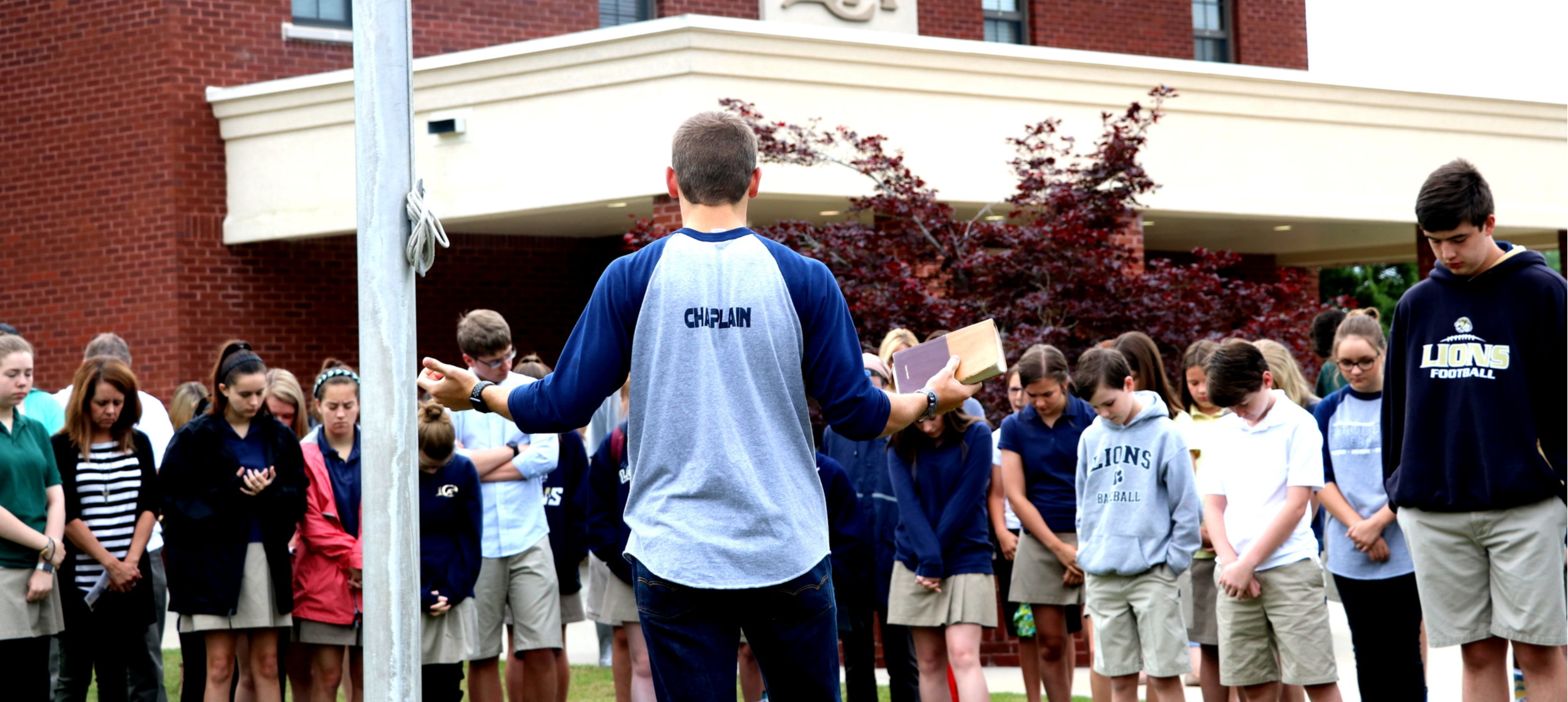 A group of students stands in a circle with their heads bowed, listening to a speaker in front of a school building, with a flagpole in the foreground.