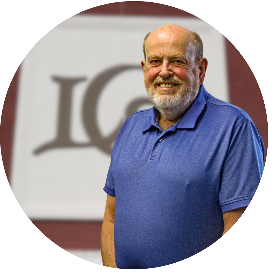 A smiling older man with a beard stands in front of a blurred background featuring a logo, wearing a blue polo shirt.