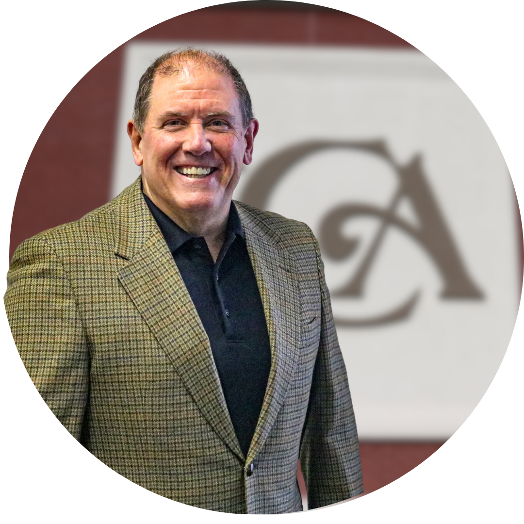 A smiling man in a checkered blazer stands in front of a blurred background featuring a logo, conveying a professional and approachable demeanor.