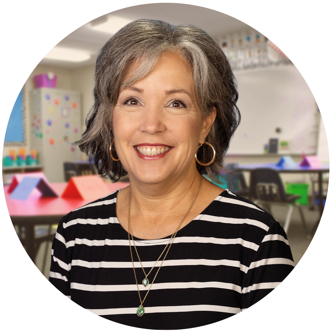 A smiling woman with gray hair wearing a striped shirt stands in a classroom setting, with colorful decorations and classroom furniture visible in the background.