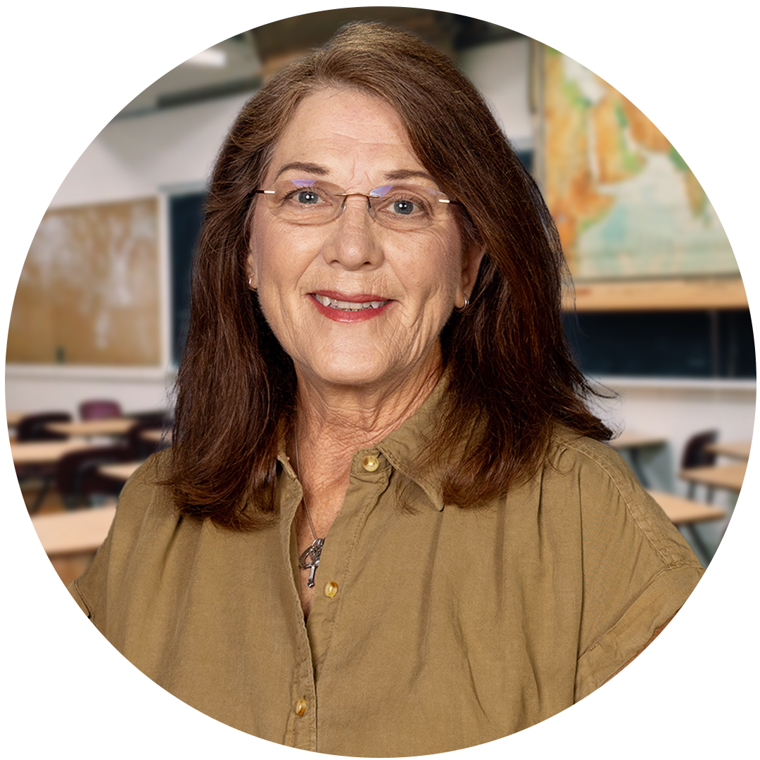 A smiling woman with glasses and brown hair, wearing a tan shirt, stands in a classroom setting with desks and a map in the background.