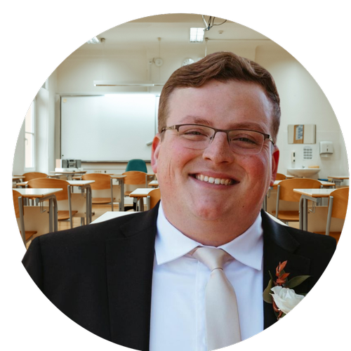 A smiling man in a suit with a white tie and a flower boutonniere stands in a classroom setting, with empty desks and a whiteboard in the background.