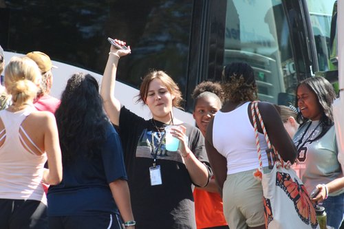 A group of people, mostly women, are gathered around a bus, with one woman raising her hand and holding a drink, while others are engaged in conversation.