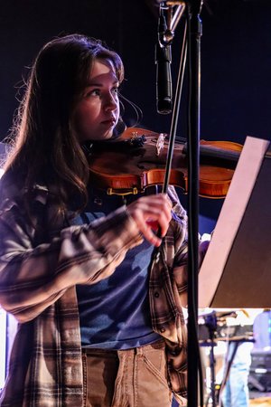 A young woman plays the violin on stage, wearing a plaid shirt and focused on her music sheet, with a microphone in the background.