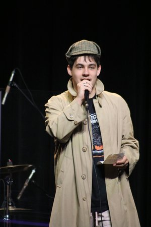 A young man wearing a beige trench coat and a newsboy cap stands on stage, holding a microphone and speaking to an audience.