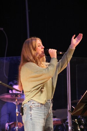 A young woman with long hair sings into a microphone while standing on stage, with a drummer visible in the background.