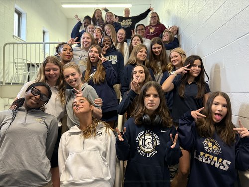 A large group of young women poses playfully on a staircase, wearing matching team sweatshirts and making fun gestures.