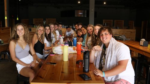 A group of young adults sitting together at a long wooden table in a casual dining setting, smiling and enjoying their time.