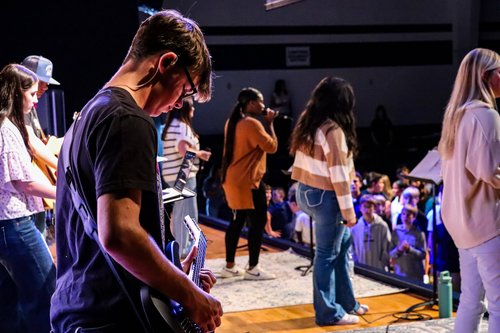 A group of musicians perform on stage, with a guitarist in the foreground and vocalists behind, while an audience watches from below.