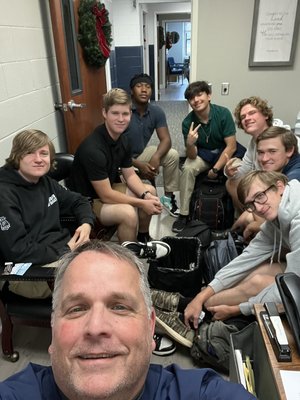 A group of eight individuals, including a smiling man in the foreground taking a selfie, are gathered in a room with a festive wreath on the door, sitting on chairs and the floor, with backpacks and equipment around them.