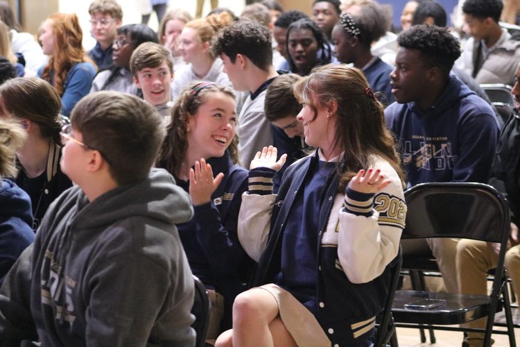 A group of students sitting in a crowded auditorium, with two girls in the foreground engaged in conversation and smiling at each other.